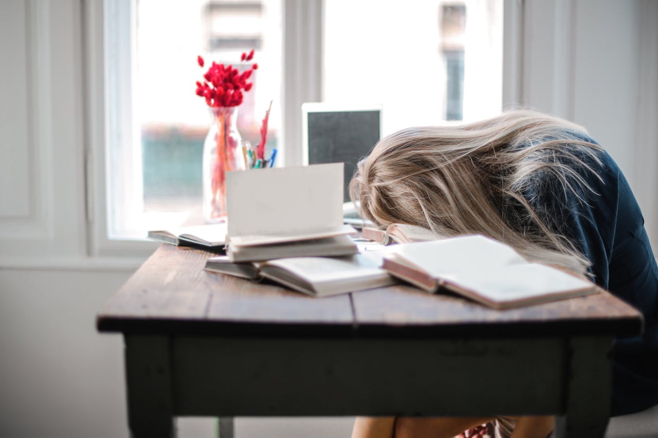 about-01 Blonde woman slumped over desk filled with books, showcasing study fatigue indoors.