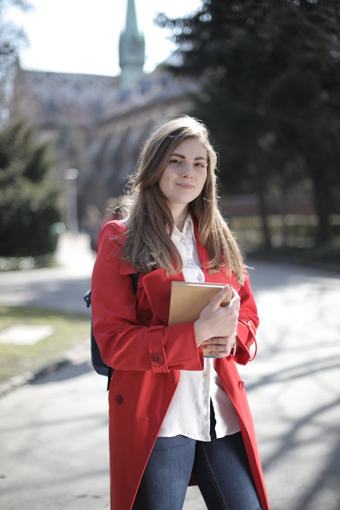 gallery-4 Young woman in a red coat smiling while holding a book outside on a sunny day.